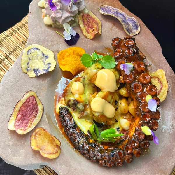 Peruvian grilled octopus with sauce, corn, edible flowers, and colorful root vegetable chips on a stone plate.