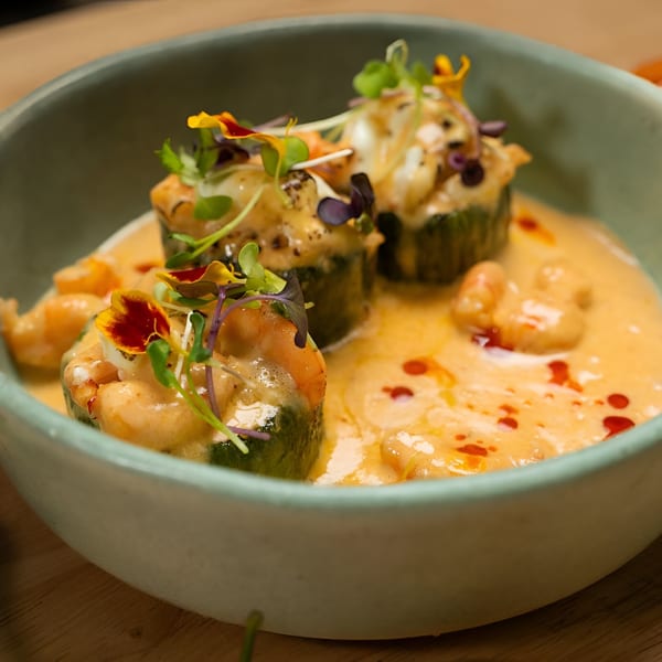 Peruvian dish with zucchini rolls, shrimp, creamy orange sauce, and edible flowers in a green bowl.